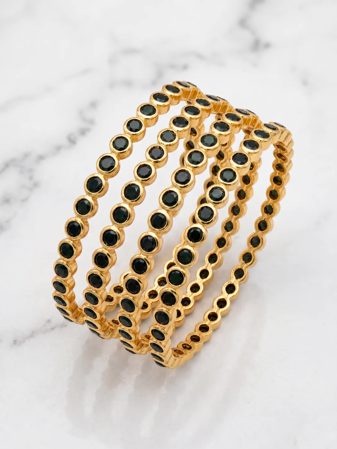 Four gold-tone bangles with round black accents arranged on a pale marble-like surface