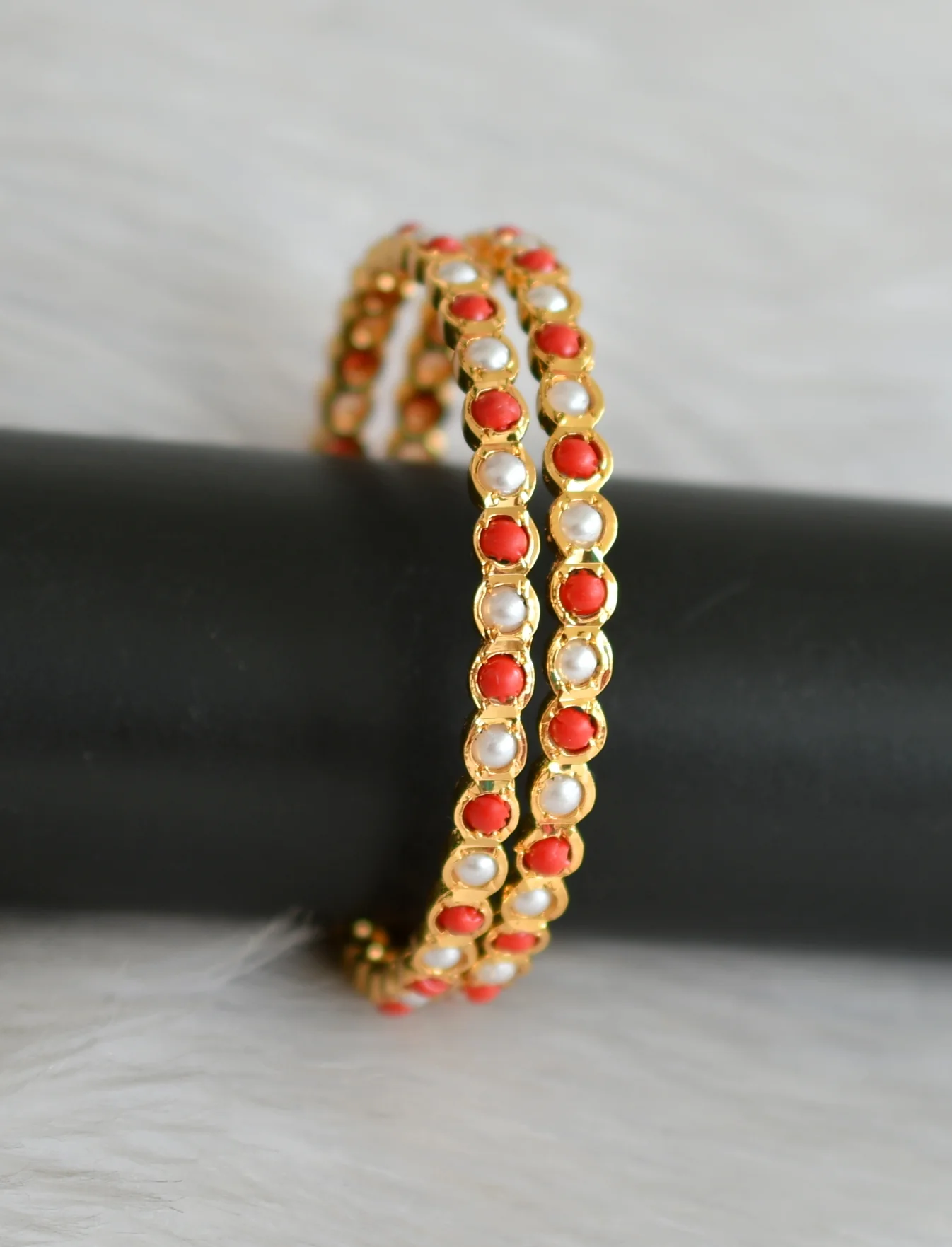 Pair of gold-tone bangles with alternating pearl-like and coral-coloured round accents displayed upright