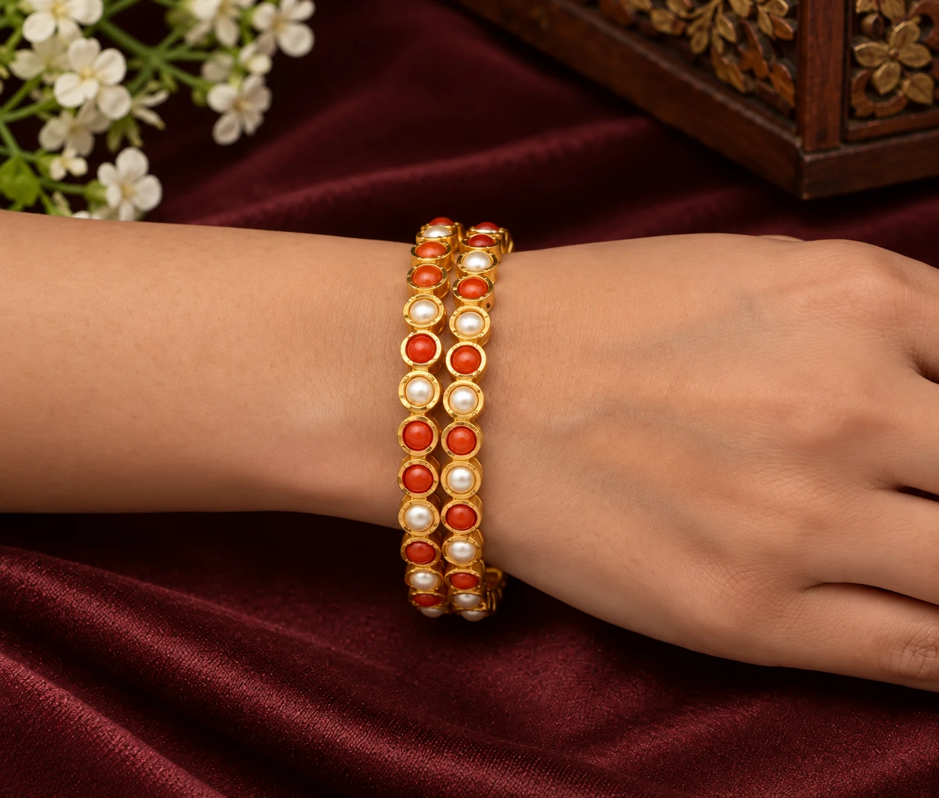 Pair of gold-tone bangles with alternating pearl-like and coral-orange round accents worn on a wrist against maroon fabric