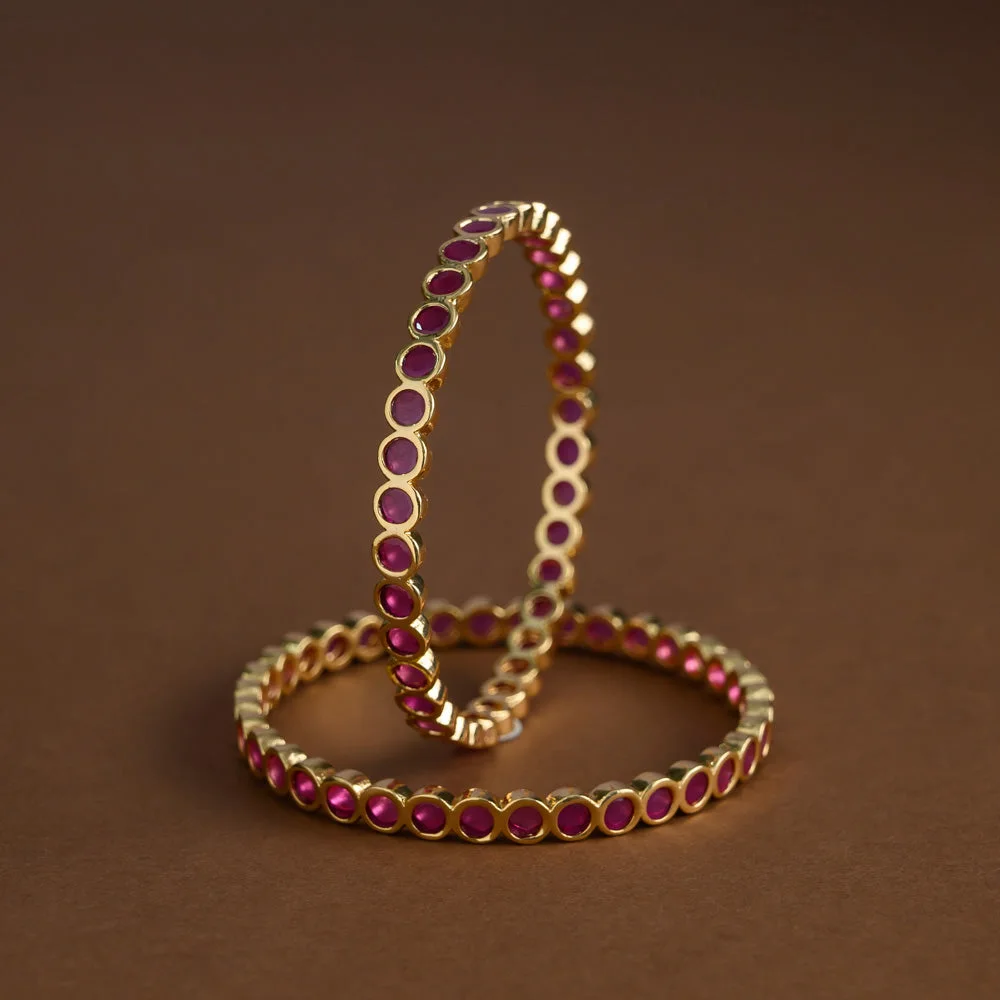 Pair of gold-tone bangles with round ruby-coloured accents arranged on a warm brown backdrop