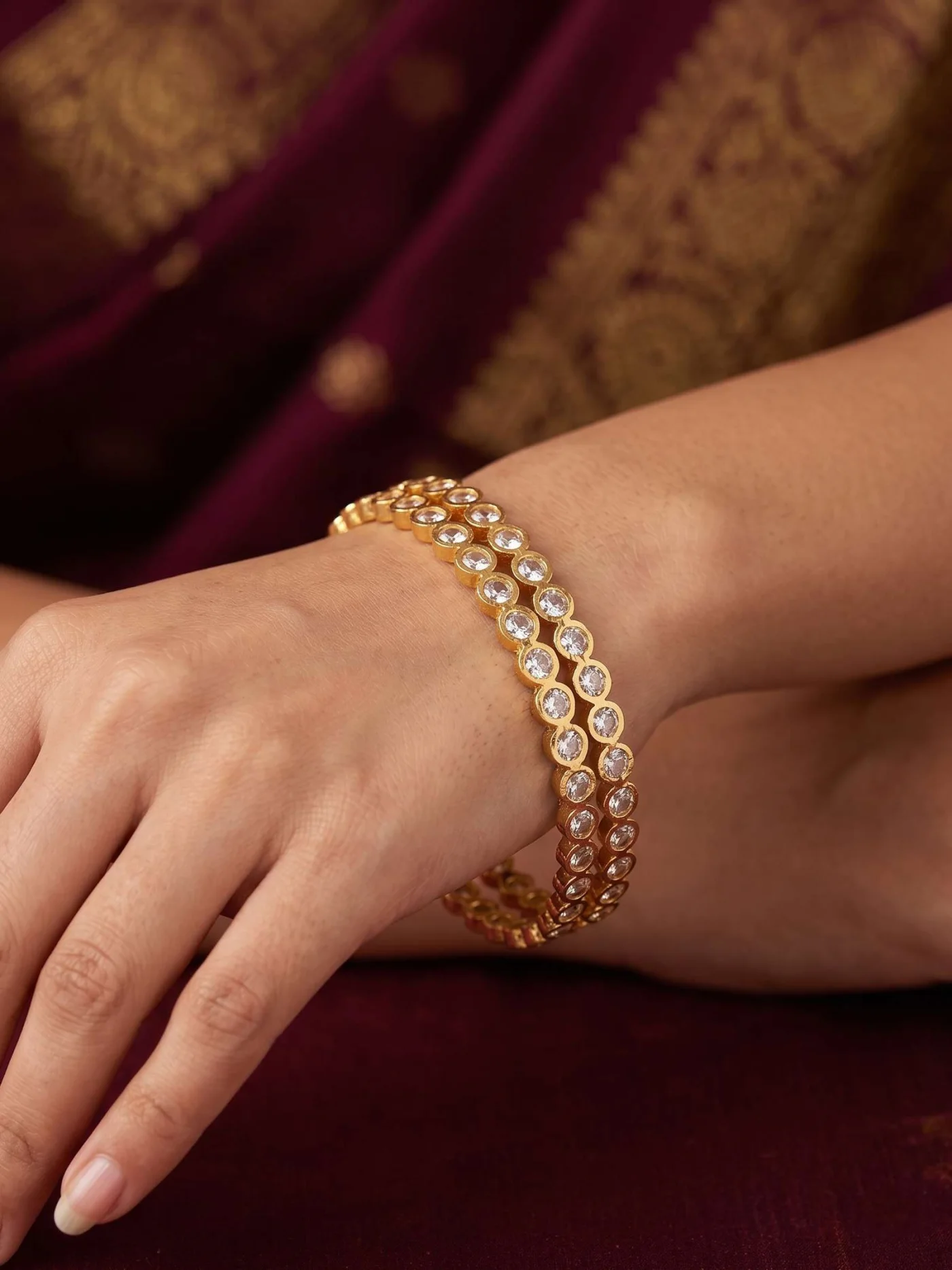 Styled wrist view of two gold-tone white-stone bangles with maroon fabric in the background